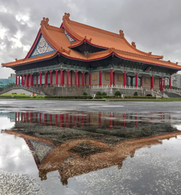The exterior of Taipei's National Concert Hall, surrounded by standing puddles of fallen rain that capture the building's reflection, and with more storm clouds in the background.
